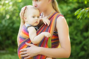 Baby Wearing Woman holding a baby in a colorful sling outdoors, gently kissing the baby's head, with green foliage in the background—capturing the tender bond after a Bloomington home birth.
