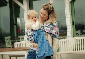 Baby Wearing A woman smiles while holding a toddler in a blue patterned baby sling outdoors near benches and a building with glass doors, reflecting the warmth often seen in St. Paul midwives supporting families.