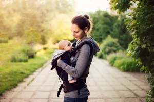 Baby Wearing Woman standing on a stone path outdoors holds a baby in a front-facing carrier, both dressed in casual hoodies. The sunlit, green setting reflects the calm support offered by St. Paul midwives for families on their parenting journey.