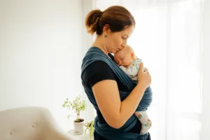 Baby Wearing A Minneapolis midwife supports a woman holding a sleeping baby in a blue wrap carrier, standing indoors near a window with sunlight and a small plant in the background.
