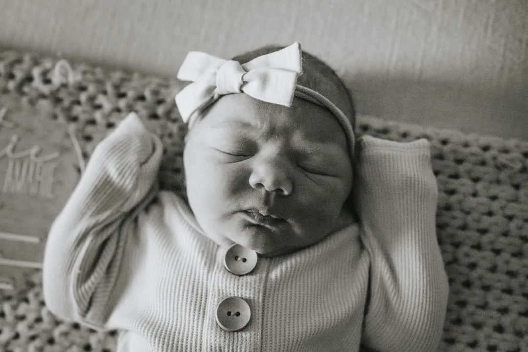 Black and white photo of a newborn baby in a bow headband and ribbed outfit, lying with arms bent—capturing the peaceful moments after a Cottage Grove out of hospital birth.