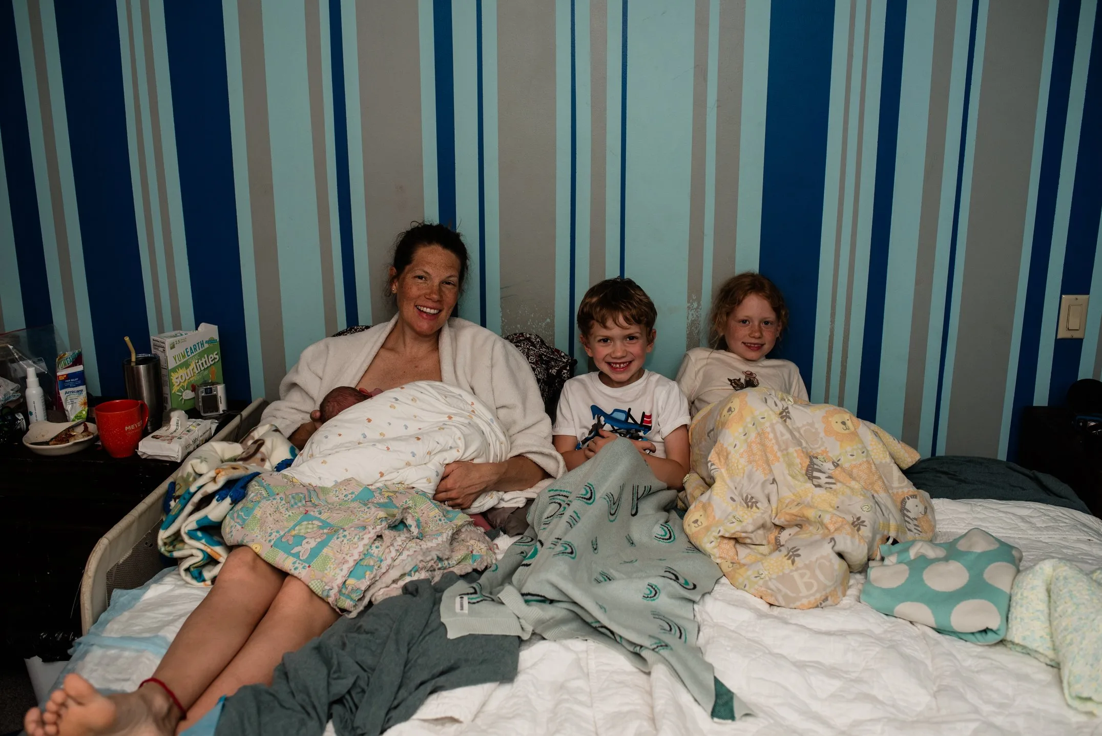 An adult and three children sit on a bed covered in patterned blankets, with a striped blue and gray wall in the background, reflecting the warmth often found in Bloomington home birth settings.