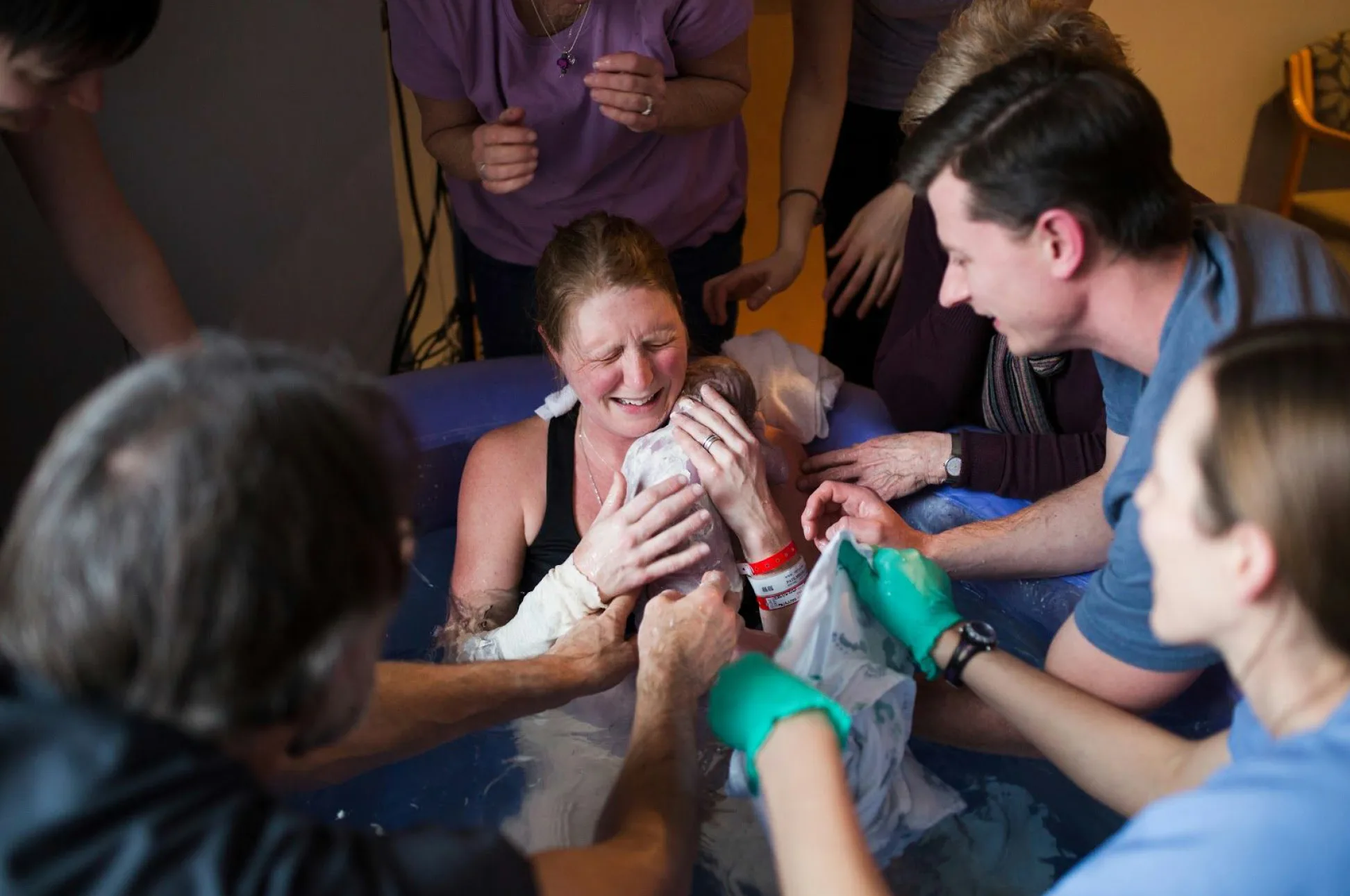 A woman in a birthing pool holds a newborn, surrounded by supportive people, including a Minneapolis midwife assisting with the water birth.