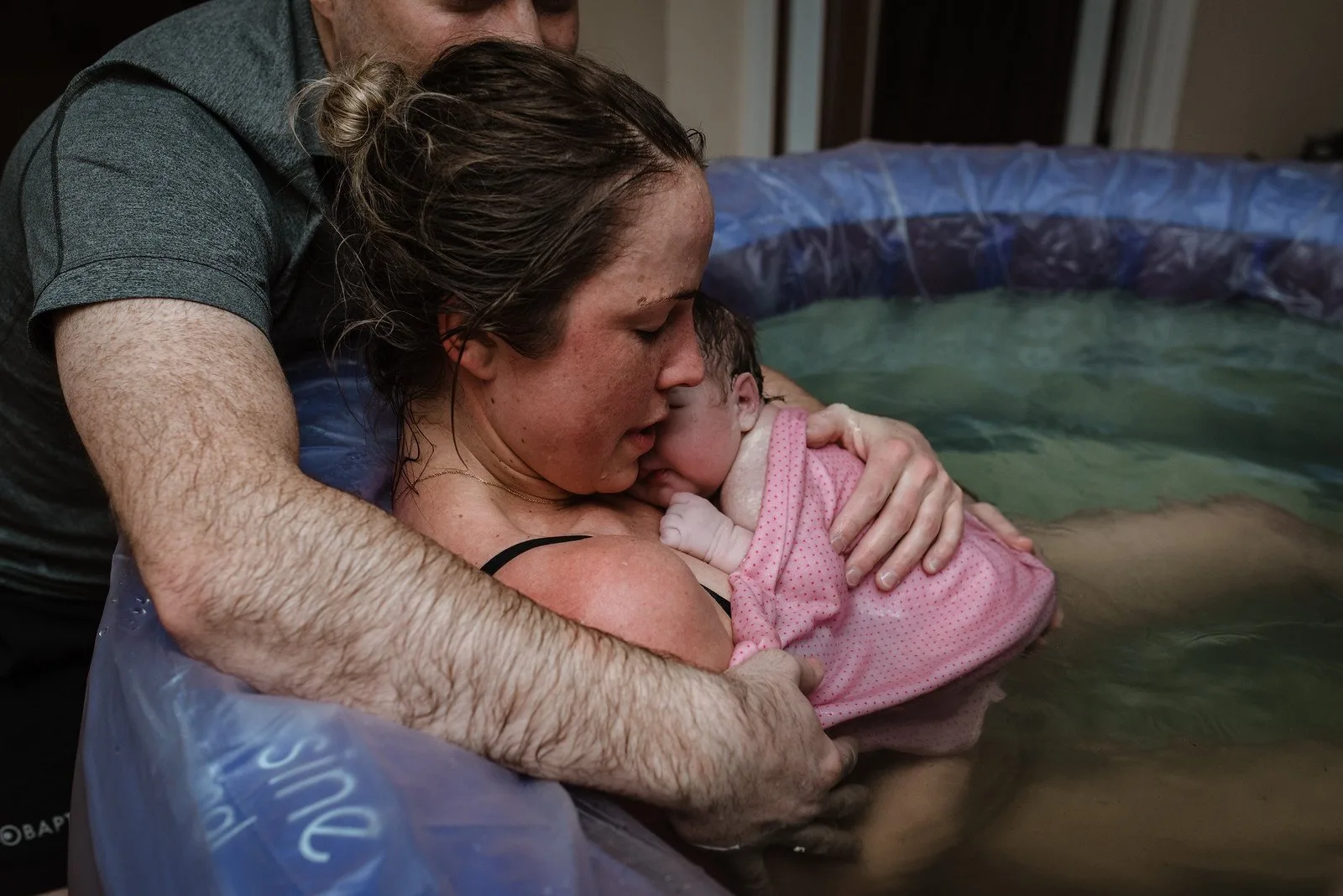 A woman holds a newborn baby wrapped in a pink blanket while sitting in a birthing pool, supported by her partner—a serene moment often guided by caring St. Paul midwives.