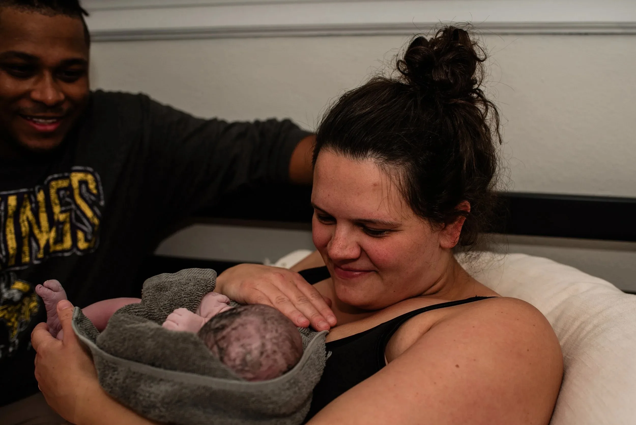 A woman holds a newborn wrapped in a towel on her chest while a man sits beside her, both appearing content after their Minneapolis midwife–assisted birth.
