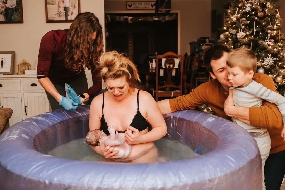 A woman sits in a birthing pool holding a newborn, with her supportive partner and child nearby, as a birth assistant—one of the caring St. Paul midwives—stands beside the pool.