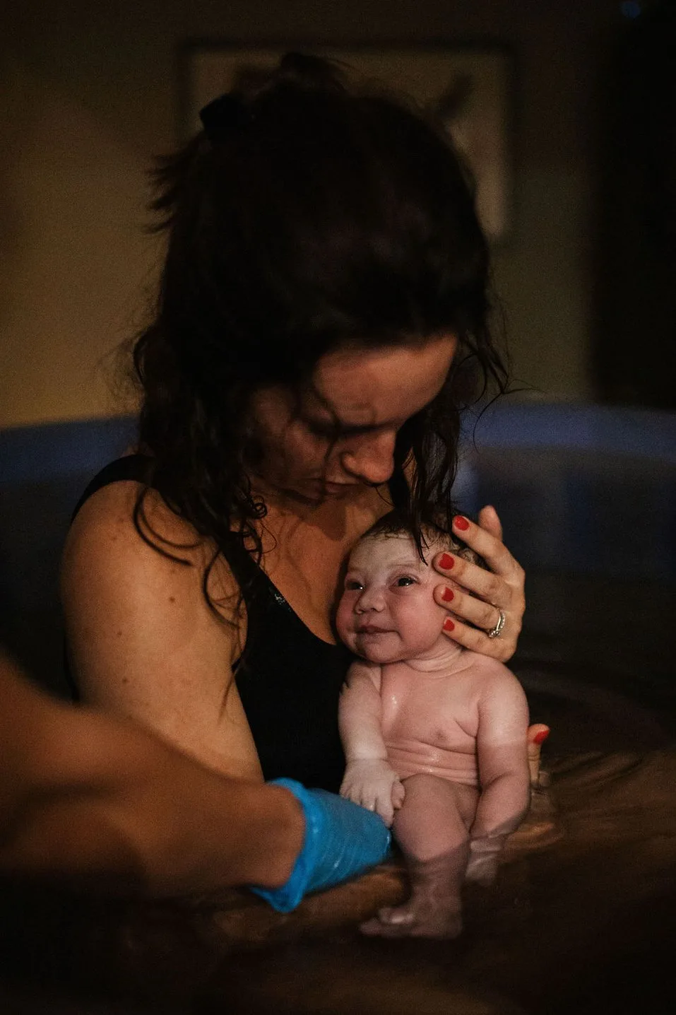 A woman holds a newborn baby in a birthing pool as a Minneapolis midwife with a gloved hand assists during a peaceful water birth.