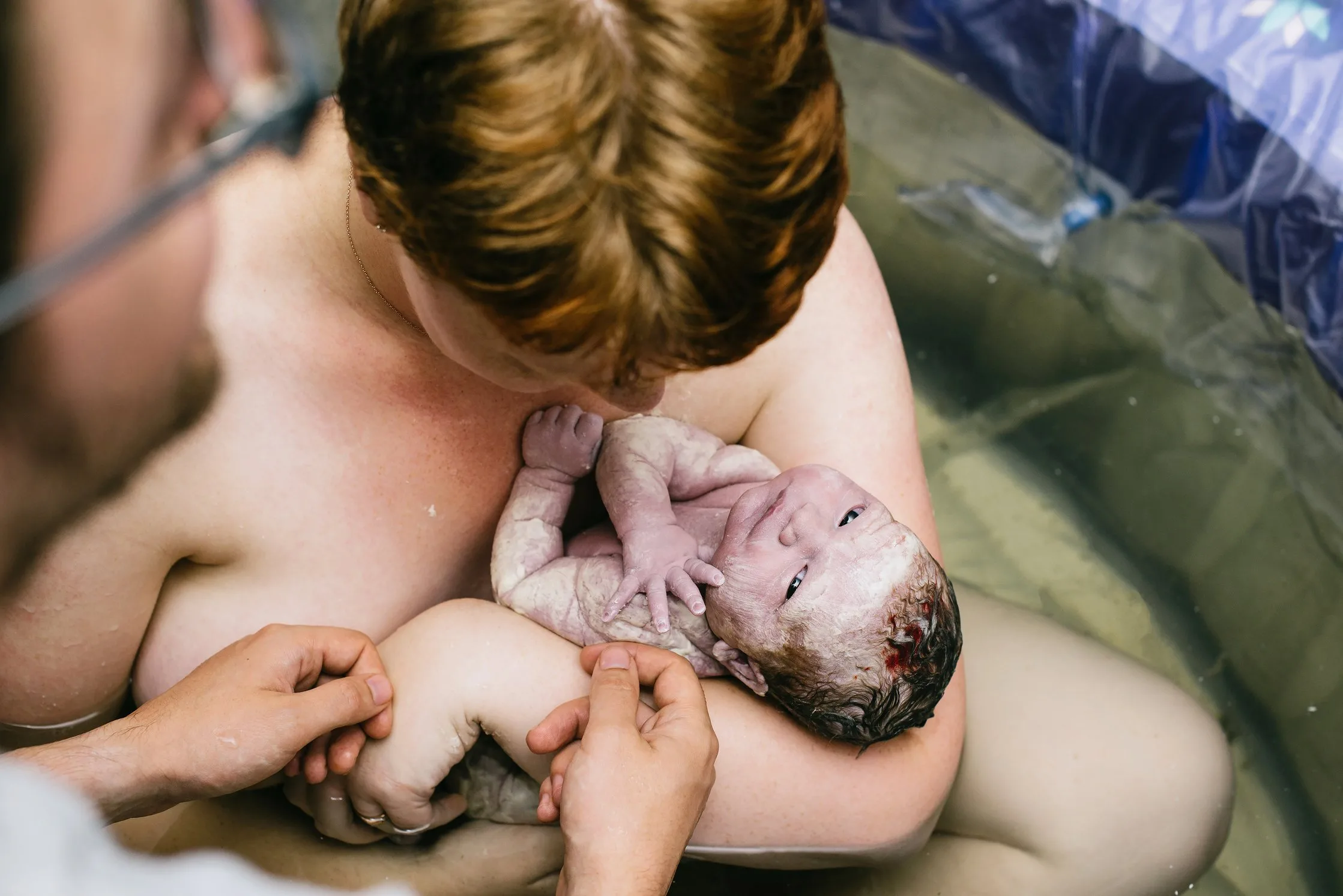 A newborn baby is held by a woman in a birthing pool while another person holds the baby’s hand, capturing the warmth of a White Bear Lake midwifery experience.