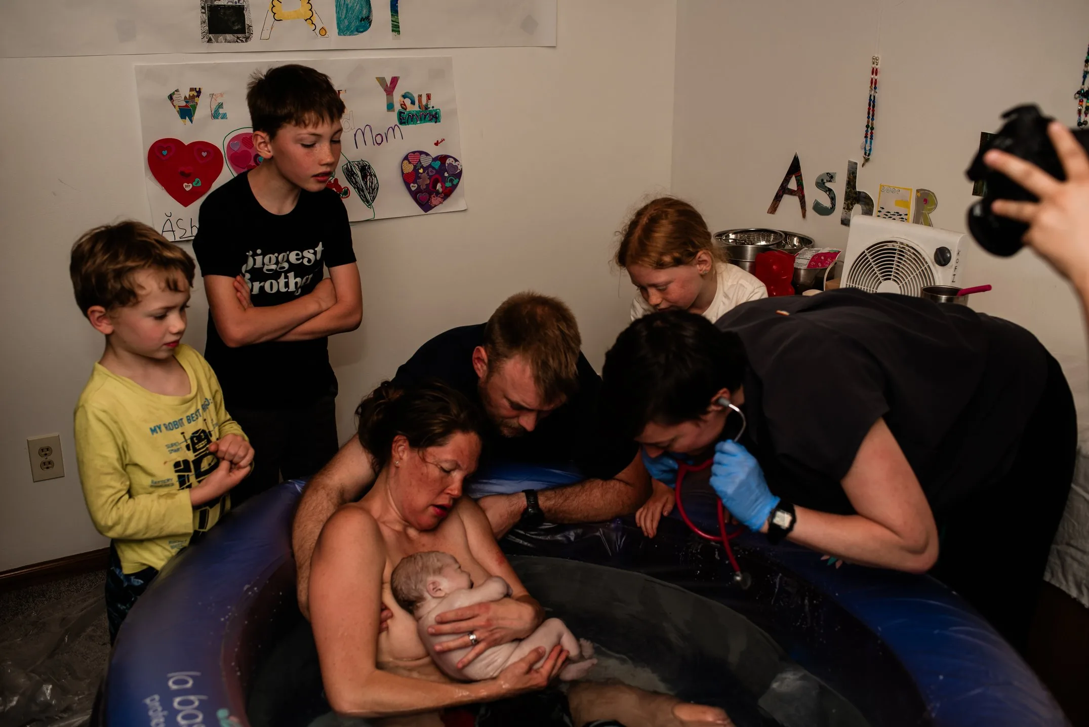 A woman holds a newborn baby in a birthing pool, surrounded by her family and a medical professional during a Bloomington home birth.